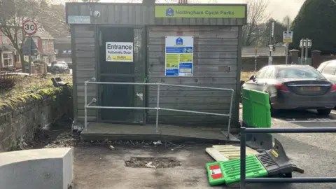 A shallow hole in a car park front of a wooden cycle storage shed