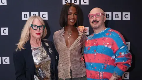 Getty Images Three people are standing on the red carpet in front of a navy board which has the BBC logo printed across it multiple times. To the left is a woman with blonde hair and red lipstick. She is wearing dark green cat-eye glasses and a gold sparkly top with a navy pinstripe blazer over it. There are silver diamond pins on her lapel. In the middle is Leomie, smiling straight at the camera. She is the tallest and is wearing a brown snakeskin shirt. She has a silver necklace on and a short black bob. To the right is a man with a flicked up moustache and beard. He has pink square-frame glasses and is wearing a bubblegum pink and blue striped jumper with silver sequins stitched onto it.