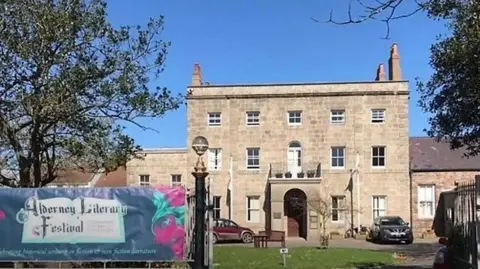 A three-story house made of light brown brick. There are two cars parked in front of the house. There is a banner on the gate to the house which reads "Alderney Literary Festival".