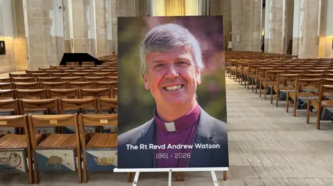 A photo of a bishop propped on a stand, captioned with "The Rt Revd Andrew Watson 1961-2026" at the bottom. It is placed inside a cathedral building lined with seats.