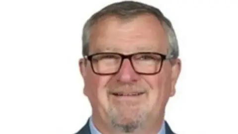 Staffordshire County Council A headshot of a man with grey hair and grey beard. He has glasses and a shirt collar and jacket can just about be seen