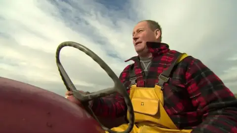 A close-up, low-angle photograph of Michael Wilson in yellow waterproof overalls and a red-and-black checked jacket sitting at the wheel of an old red tractor. The large steering wheel is prominent in the foreground, and the scene is set outdoors beneath a bright, cloud-filled sky.