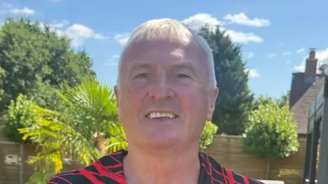 Clive Seeley A photo of a man with short white hair smiling. He is in his garden. It is a close up photo and you can see the top of his black and red football shirt. 