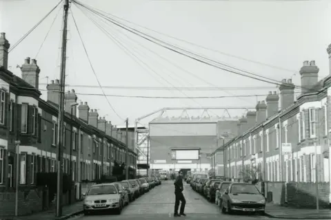 Jason Scott Tilley/Lower Block A black and white image of a terraced street, with cars parked either side. At the end is Highfield Road stadium. A man can be seen crossing the road. 