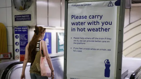 EPA A woman with red hair carries a pink tote bag as she travels down an escalotor at a Tube station in London. Next to here is a TFL notice that reads 'please carry water with you in the hot weather'.