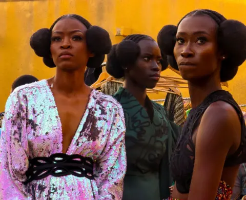 Getty Images Three models with their hair styled in afro puffs looking on backstage at a fashion show.
