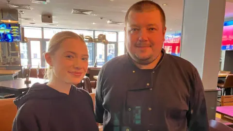 Seb Cheer/BBC A woman and man stand smiling at the camera. They are in a pub with TV screens in the background.