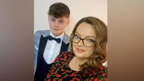 Kerryann Barr A teenage boy and woman pose for a selfie. He is wearing a suit, dickie bow and while shirt, and blue checked jacket. To his left a woman stands holding the phone to take the picture. She is wearing a red dress and has brown shoulder length hair