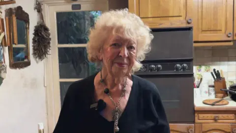 A woman stands in a kitchen of her cottage smiling at the camera. She has curly white hair and is wearing a large necklace and a black top.