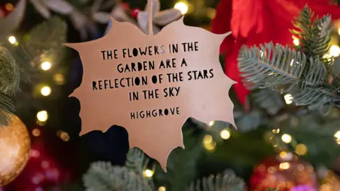 PA A bronze leaf shaped Christmas tree decoration hangs from a branch, surrounded by fairy lights and gold and red decorations. The leaf reads: "The flowers in the garden are a reflection of the stars in the sky. Highgrove."