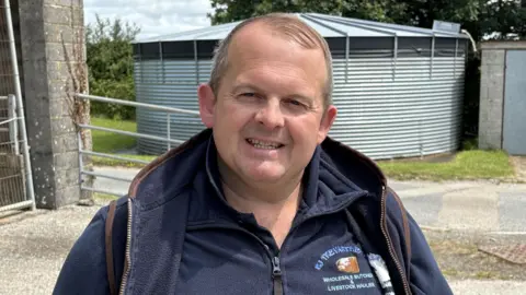 A man with greying light hair, smiling, while wearing a blue fleece jacket standing in front of a corrugated metal silo. 