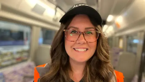A woman with long brown hair, glasses and a black cap is stood inside a train carriage.