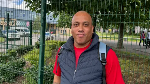 A bald man wearing a red short sleeved t-shirt and navy blue bodywarmer. He is wearing a rucksack and stood in front of a metal fence