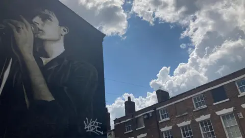 The Ian Curtis mural in Macclesfield, which is of a man holding a microphone on the side of a building, with blue sky overhead and some clouds.