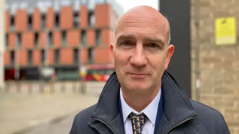 Peter Dervin has a shaved head and is wearing a black jacket over a white shirt and a navy and golden tie. He is offering a resigned smile to the camera. Behind him are buildings and a bus going past.