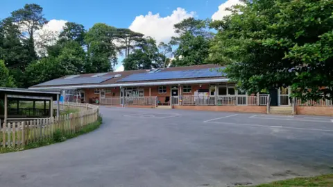 Nettlebed Community School Outside image of Nettlebed Community School, which has solar panels covering its roof and is surrounded by trees and greenery