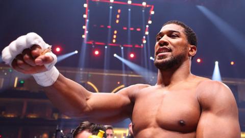 Anthony Joshua in a boxing ring smiling and raising his right fist towards the camera