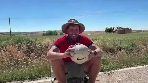 James sits on his bag amid high temperatures as he is interviewed while holding his football. He has a large sunhat on with a red T-shirt and shorts. He is unshaven. Grassland can be seen behind him and a blue, cloudless sky.