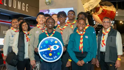 Twelve scouts including adults and children with one at front holding a large, round badge showing the design for new space badge