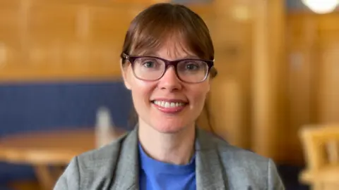 Joney Faragher has her brown hair tied back, a long fringe and glasses. She wears a grey jacket and blue top, and is smiling. Blurred chairs and tables can be seen in the background.