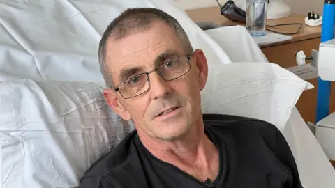 A close-up of Tommy Richards' face in a hospital bed. He has brown eyes and thin framed glasses.