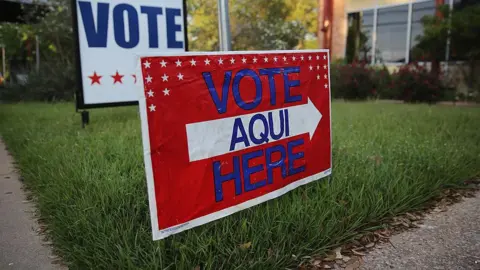 A red, white and blue lawn sign that says vote here in both English and Spanish.