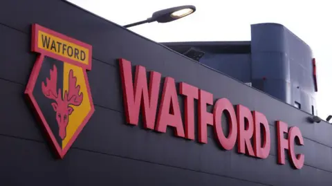 A general view outside Vicarage Road stadium. There is the black and yellow crescent of Watford which has a red moose on it. To its right are the words WATFORD FC in red. 