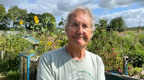 A man wearing a green t-shirt and glasses looks at the camera, standing in front of an allotment with sunflowers on a community farm