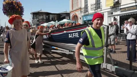 Women wearing cream aprons and orange mob caps are pulling and pushing a boat called Alfie through the streets. Spectators are applauding them.