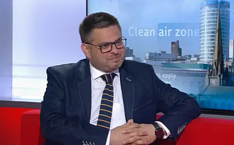 A man in a suit sitting on a red sofa in a TV studio