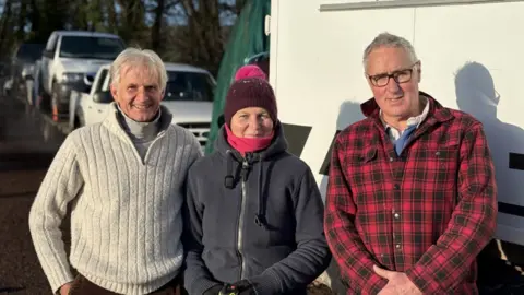 Three people wrapped up on a cold, bright day in front of a convoy of mainly white vehicles, some on trailers