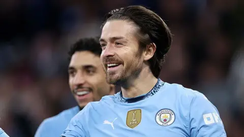 Getty Images Jack Grealish smiles on the pitch wearing a Manchester City shirt. His hair is held back by a headband 