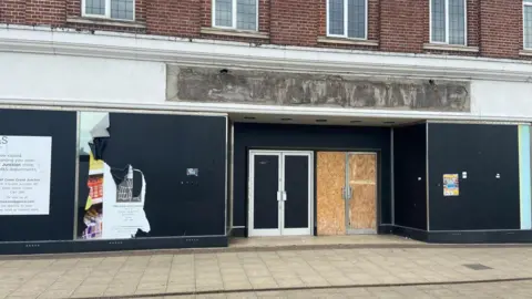 A brick-built, two-storey building with blacked-out windows and doors. There are posters and damage to some parts of the black cladding.