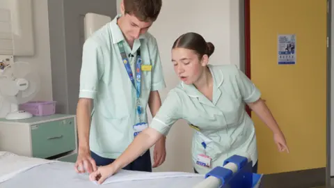 Two hospital workers, a man and a woman, are pictured making a bed in a hospital room. They are wearing light green uniforms and lanyards.