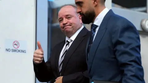 A man in a black suit and tie, giving a thumbs up to the camera, with another mad standing by his side