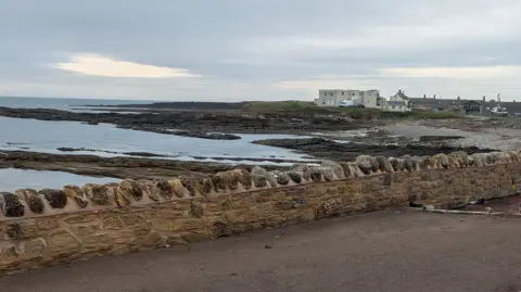 The coastal defence wall along Harbour Road in Beadnell, Northumberland. The stone wall is built near the shore. The sea can be seen behind it. A village can be seen in the distance.
