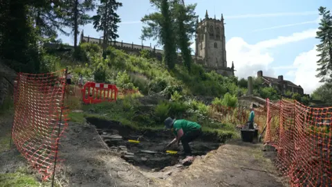 A man in a green t-shirt and wide brimmed hat is working at an excavated archaeological trench on a slope below a church.