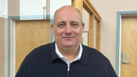 BBC There is a man with a dark,navy-blue jacket which is slightly zipped up. He has a white shirt on underneath it. He has grey receeding hair and stubble facial hair.