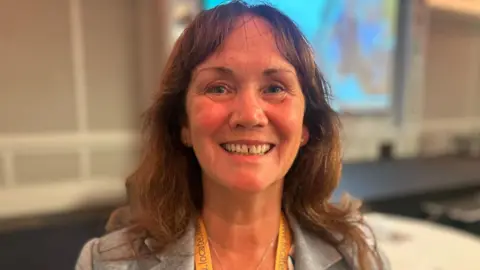 BBC Diane Kelsey MLC smiles at the conference. She has long brown hair and wears a grey suit jacket.