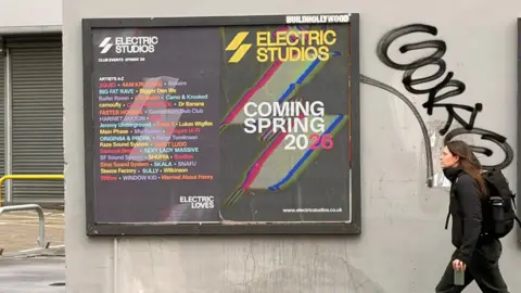 Dan Green/BBC A huge billboard gig poster against a wall. A commuter in black with a large bag walks past.