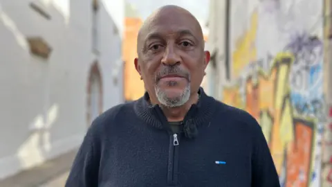 Desmond Brown stands in an alley next to a wall covered in street art. He is wearing a navy blue half zip fleece. He is bald and has a grey and black goatee.