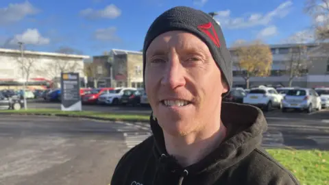 BBC Steve Bunn looks into the camera, wearing a woolly hat and hoodie in front of the leisure centre car park.