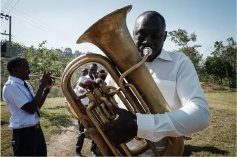 AFP Man wearing white shirt blowing a brass horn outside.