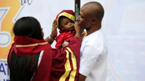 EPA A graduating student wears her cap for her child outside the venue during 53rd convocation ceremony of the University of Lagos, Nigeria, 18 January 2023. The University of Lagos, one of the first generation universities in Nigeria, celebrates its 60th anniversary with 53rd convocation ceremony graduating two octogenarian students for graduate studies.