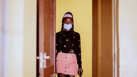 AFP A contestant at Miss Freedom Day seen in a hallway. She is wearing a pink mini skirt and a tiara. She has a mask on.