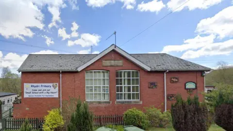 Google Rhydri Primary School pictured from the outside. There is a gate around it with bushes and trees in the foreground. The school sign can be seen on the left.