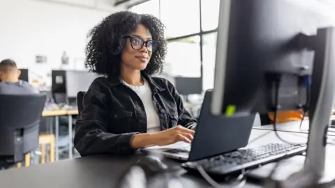 Getty Images A woman sits at her desk at work. She has a laptop and a desktop monitor