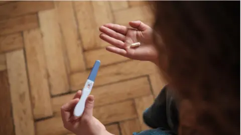 Woman holding pregnancy test and abortion pill while sitting on sofa
