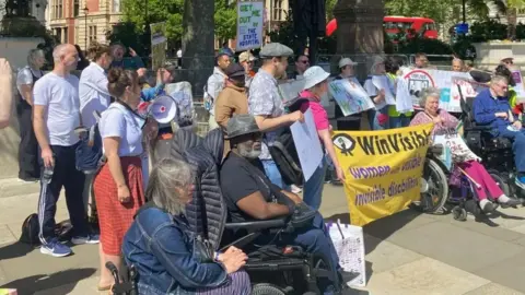 A crowd of around 30 protesters of varying ages stand in a protest. They are all looking to the left and two are holding a yellow banner reading "WinVisible". Two people in the foreground are wheelchair users. Buildings and a red London bus can be seen in the background.