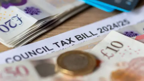 A close up shot of a council tax bill with bank notes, a pound coin and bank cards. There is a pile of £20 notes in the background.